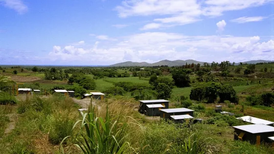 A sprawling rural landscape shows small buildings among patchworks of green fields, with distant hills under a partly cloudy sky.