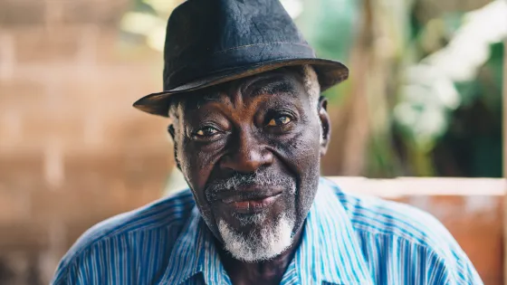 An elderly man with a warm smile wearing a black hat and a blue striped shirt.
