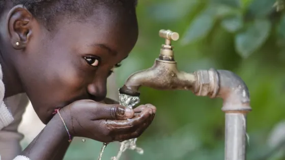 A young girl drinks water from a flowing tap, cupping her hands to catch it.