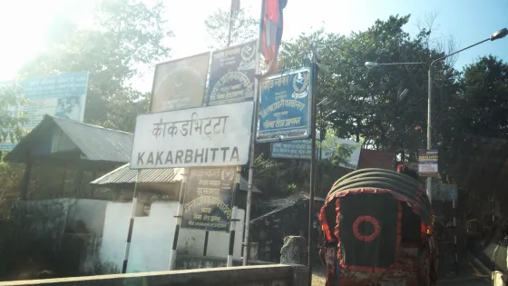 View from a vehicle of a rickshaw and multiple street signs, including one labeled "Kakarbhitta," in a sunlit, bustling street scene.