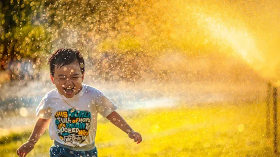 A young boy joyfully playing in sprinkling water with sunlight casting a golden glow around him.