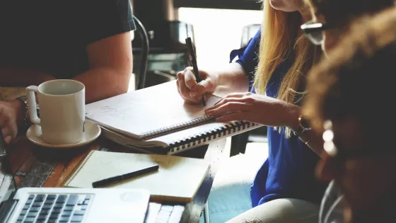 Three people sitting at a table in a café, collaborating and taking notes with laptops and notebooks visible.