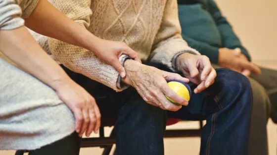 A younger person gently holds the hands of an elderly person who is sitting and holding a small ball.
