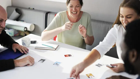 A group of colleagues laugh and engage in a discussion around a table scattered with photographs and various items.