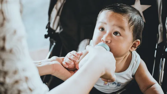 A baby holding a bottle while being fed by an adult's hand.