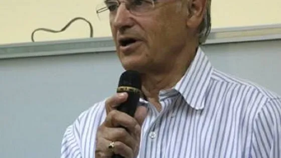 An elderly man speaking into a microphone in a room with a beige wall.