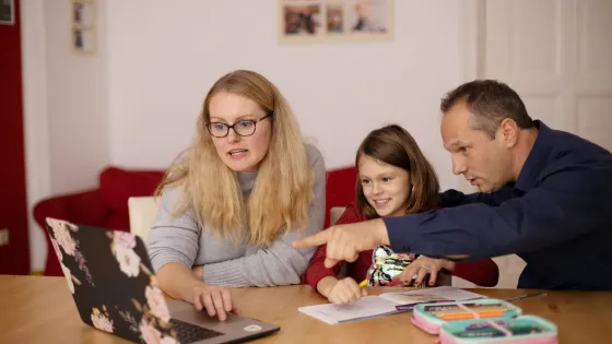 A woman, a man, and a young girl sit at a table with a laptop, engaging in a family activity together.