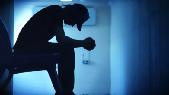 A silhouette of a person sitting with their head bowed down in a dimly lit, blue corridor.