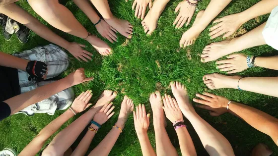 A group of people sitting in a circle on grass showing their bare feet and hands meeting in the center.