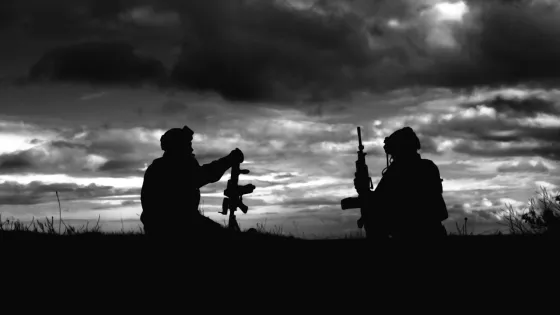 Silhouetted figures of two soldiers with equipment in a field against a cloudy sky.