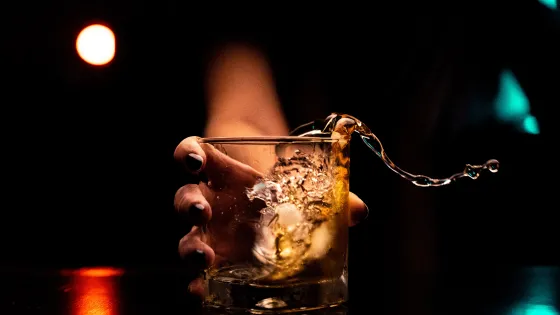 A hand tipping a glass with amber liquid, causing a splash against a dark, softly lit background.
