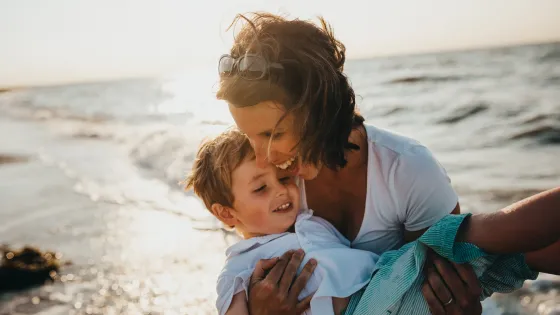 A woman tenderly embracing a young boy as they both smile near the ocean waves at sunset.