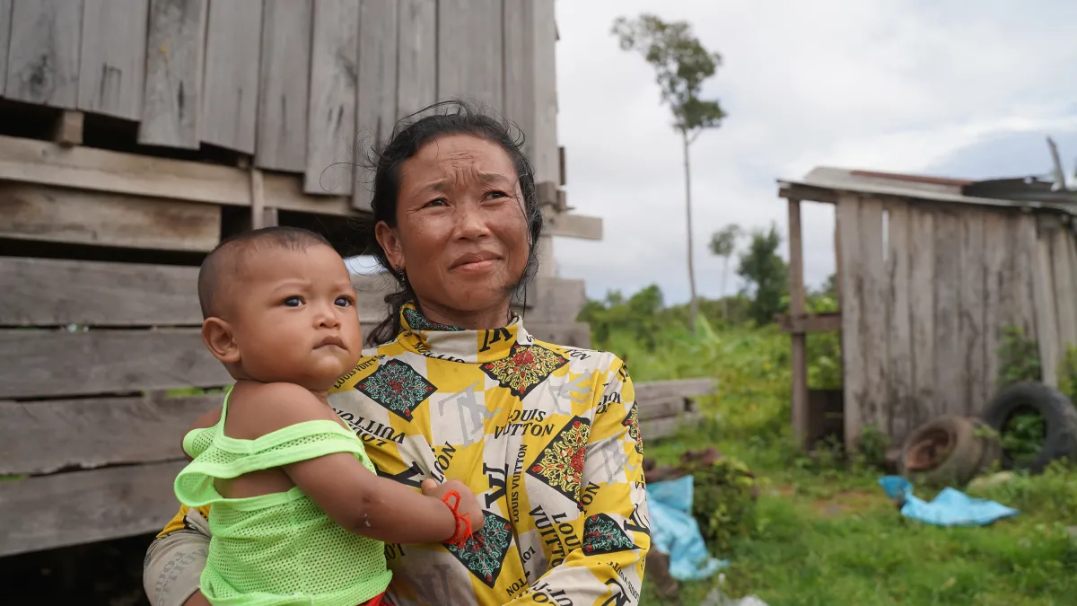 A woman holding a young child stands outdoors near a wooden structure with a rustic rural backdrop.