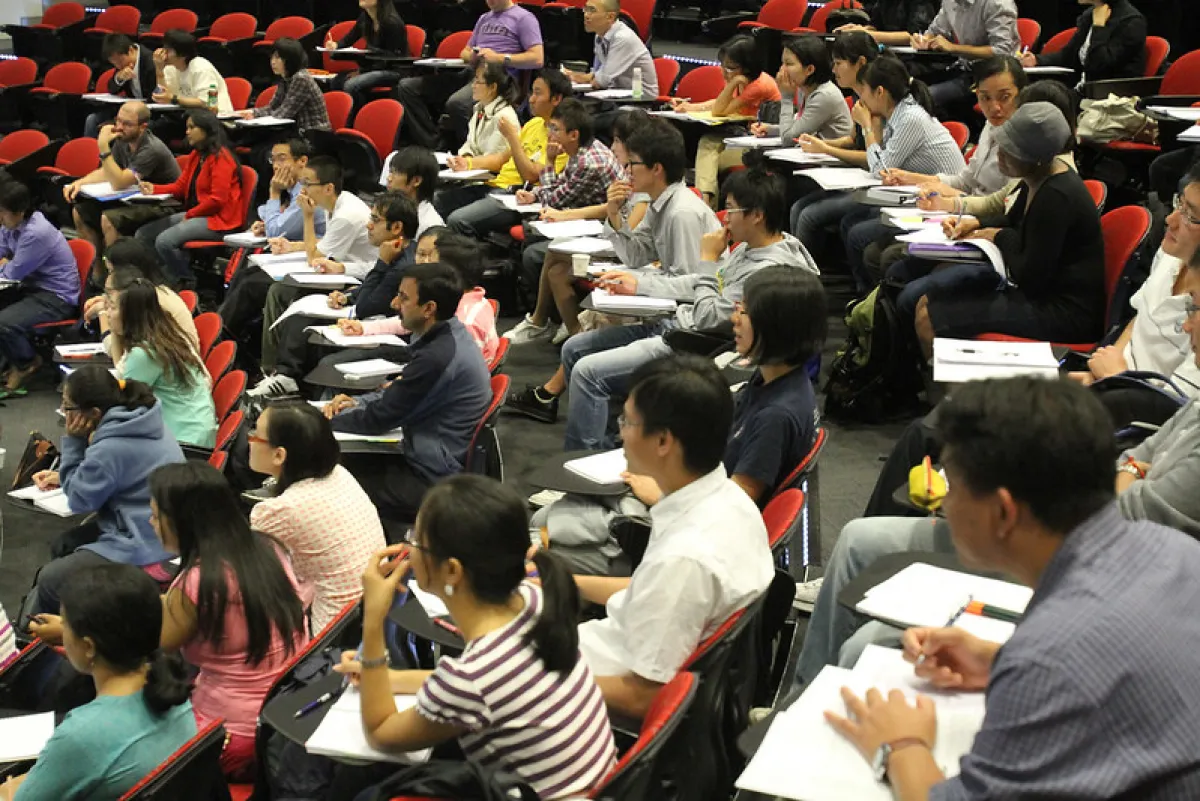 A diverse group of students sits in a university lecture hall, listening intently and taking notes.