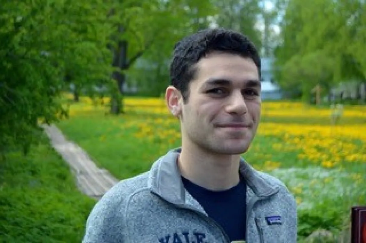 A young man smiling gently in a park with lush green trees and a field of yellow flowers in the background.