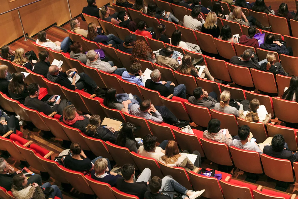 Audience members sitting in red seats in a sloped auditorium, some looking at their phones.