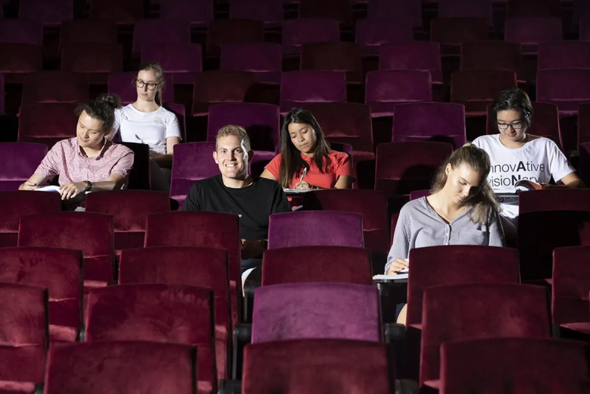 Five students are sitting apart in a dark lecture hall with purple seats, each engaging in different activities such as writing and using laptops.