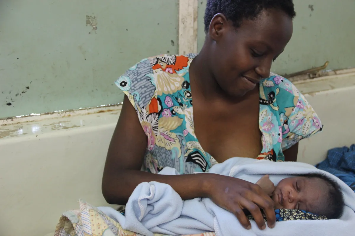 A woman smiles gently while holding a sleeping newborn baby wrapped in a light blue blanket.