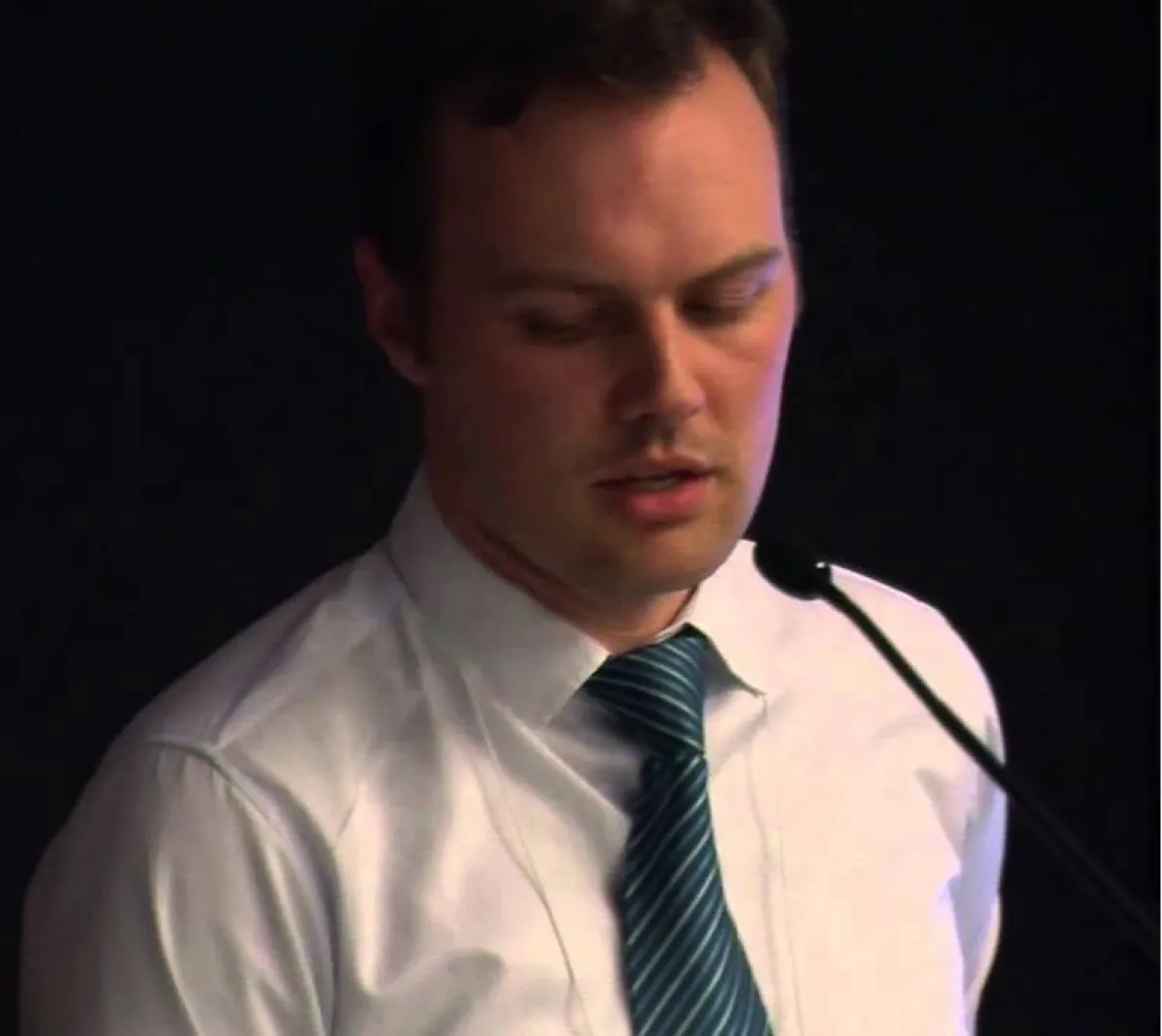 A man in a white shirt and striped tie speaks into a microphone, with his eyes closed and a focused expression.