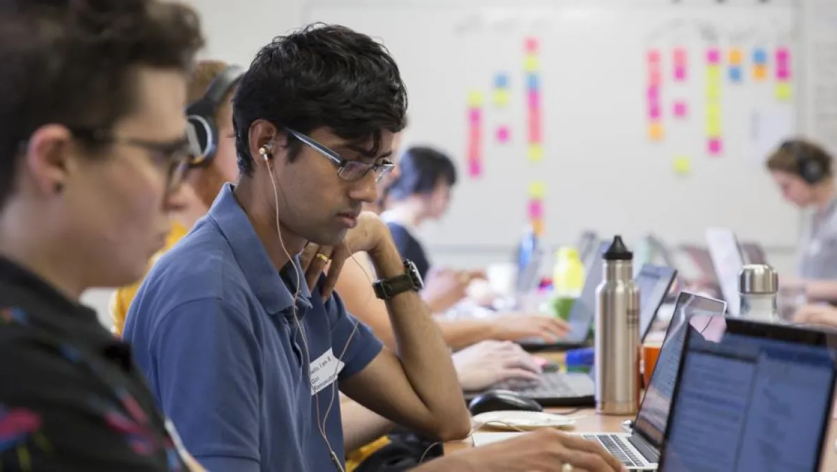 Several people wearing headphones are focused on their laptops in a busy workshop setting with notes and stationery visible on the tables.