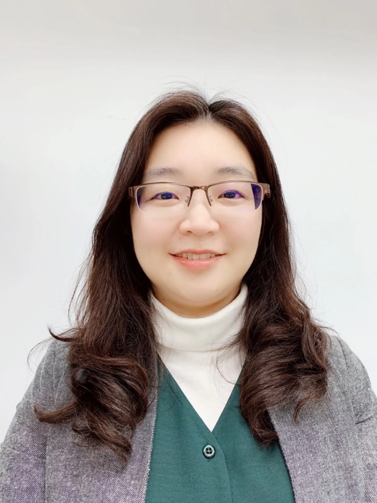 Portrait of a smiling woman with glasses, wearing a white turtleneck and a gray cardigan over a green top.
