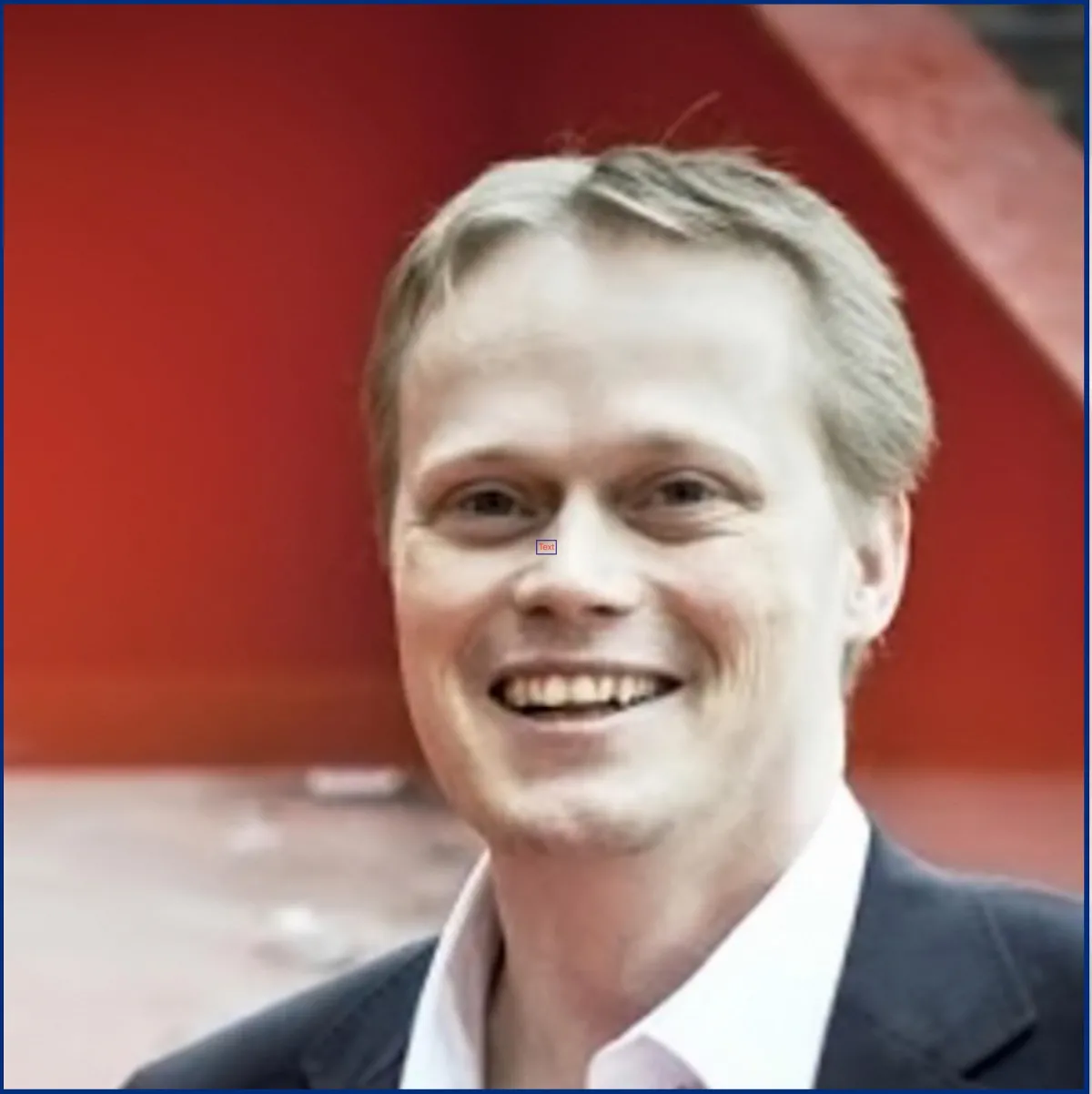 A portrait of a smiling man with blond hair, dressed in a dark suit, in front of a red background.