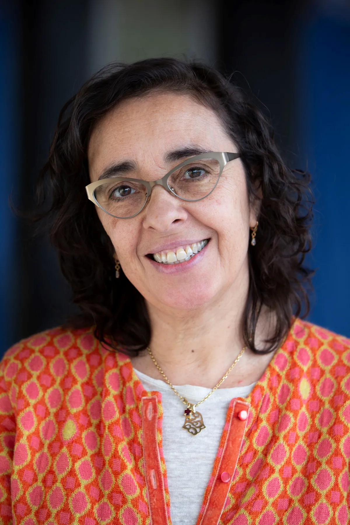 A smiling middle-aged woman wearing glasses, a vibrant orange and pink patterned jacket, and a gold necklace.