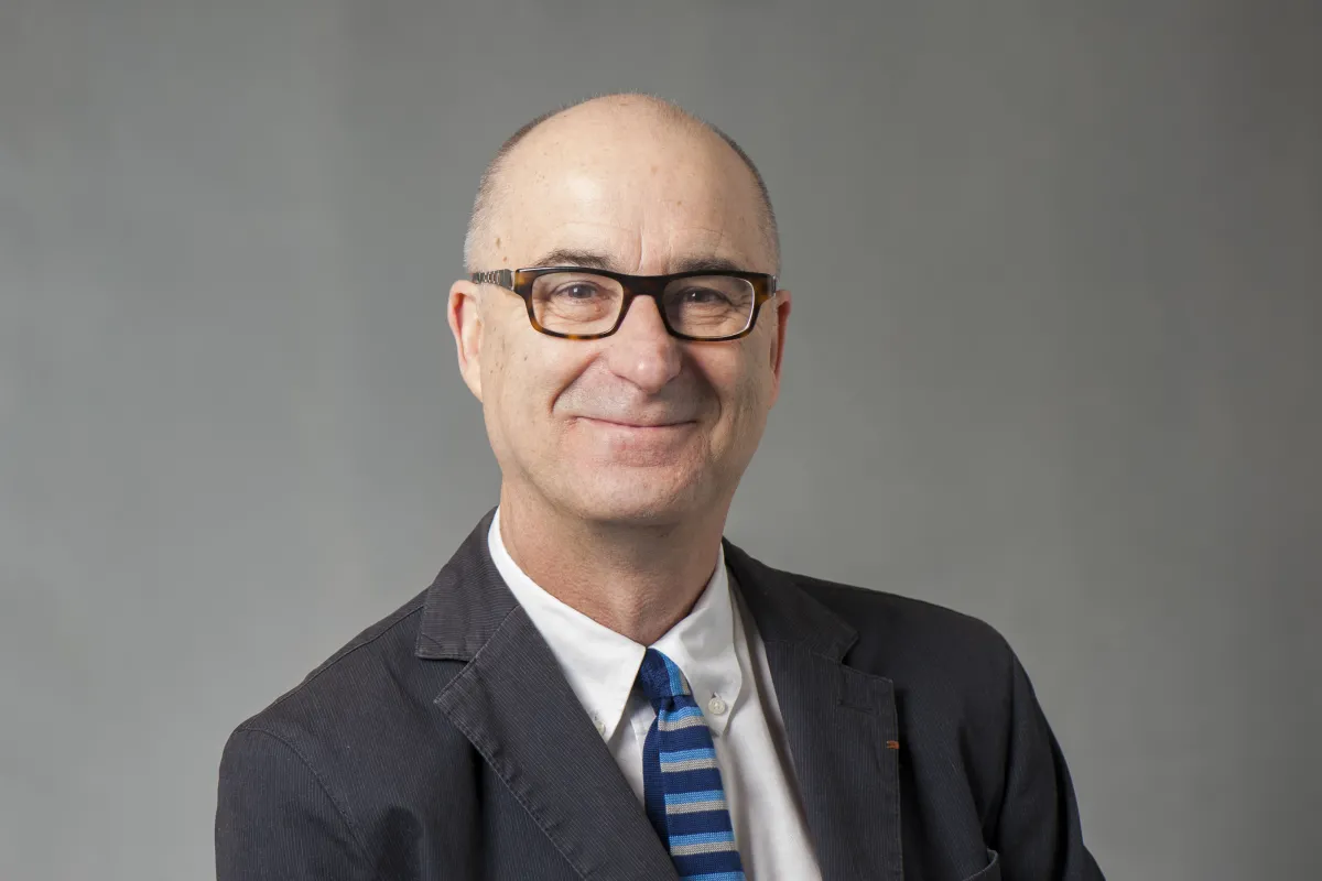 A smiling older man wearing glasses, a suit, and a striped blue tie poses against a gray background.