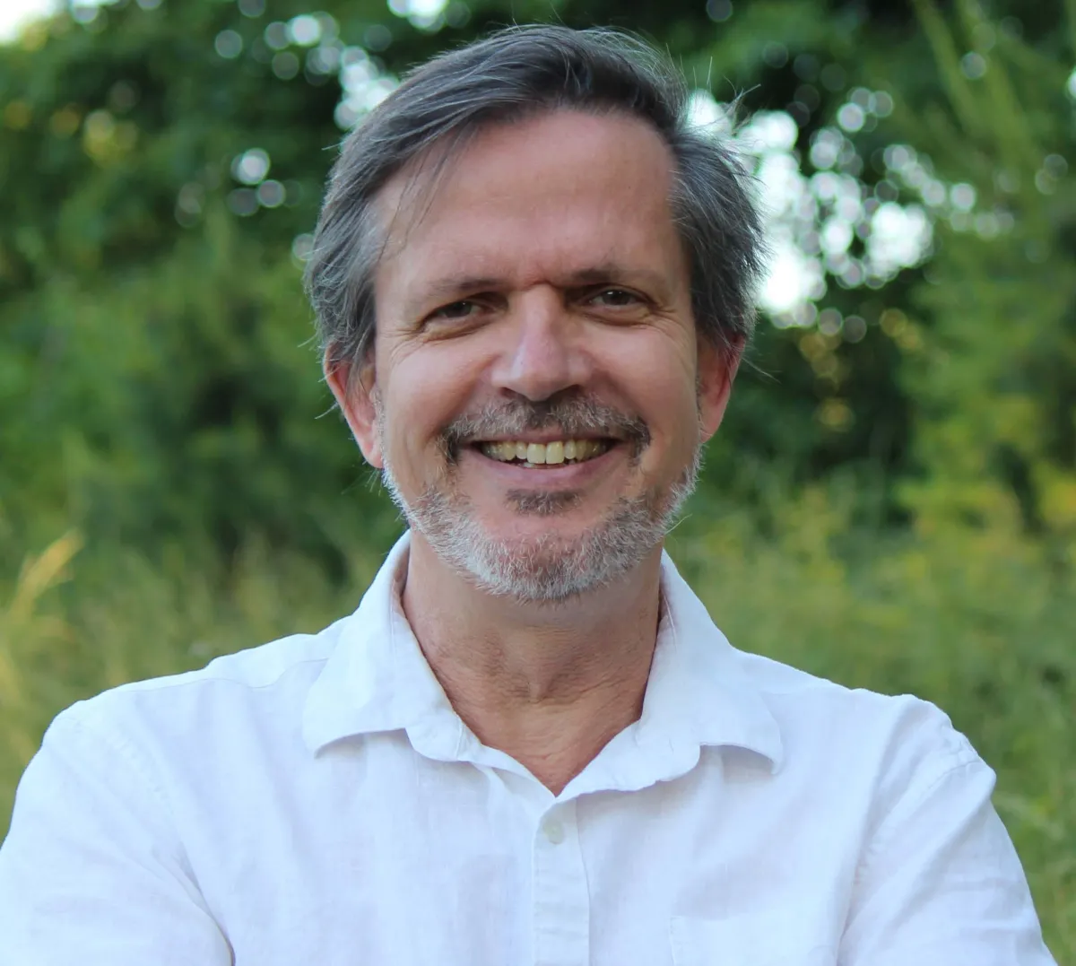 A smiling man with graying hair and a beard, wearing a white shirt, standing in front of a background of green foliage.