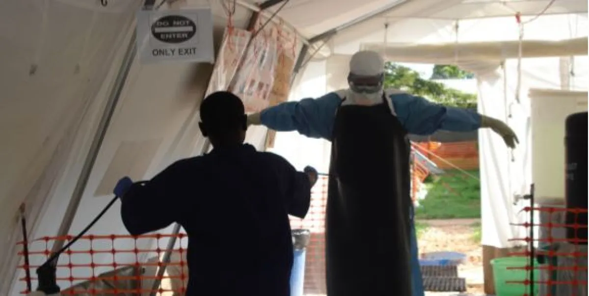 A healthcare worker in protective gear guides another person through a medical tent exit.