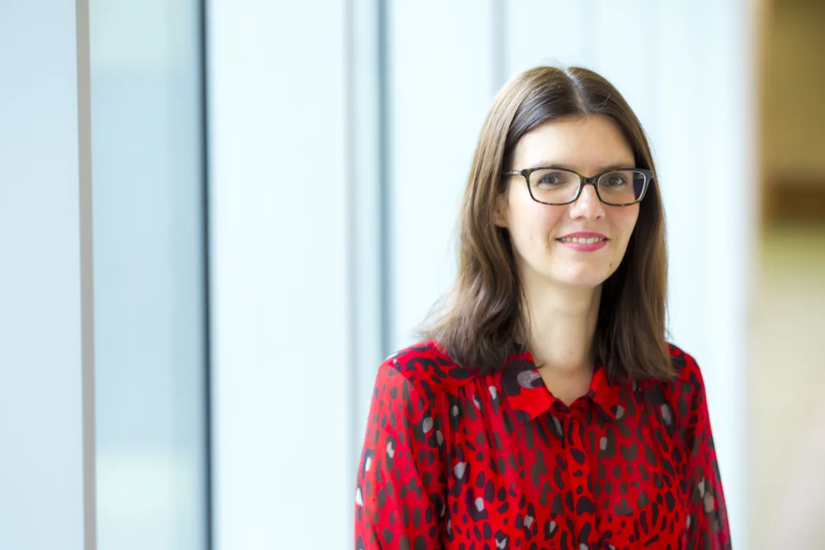 A woman in a red blouse with a black heart pattern stands smiling in a brightly lit office space.