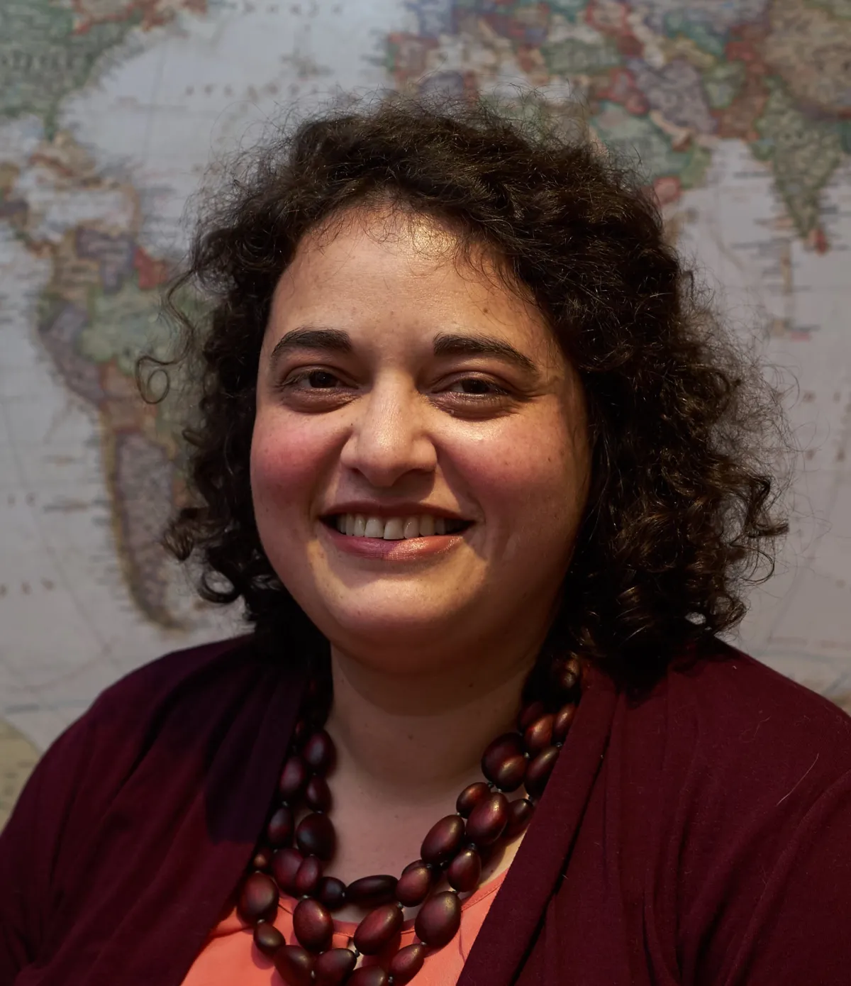 A woman smiling at the camera with a large map in the background, wearing a maroon cardigan and a chunky red necklace.
