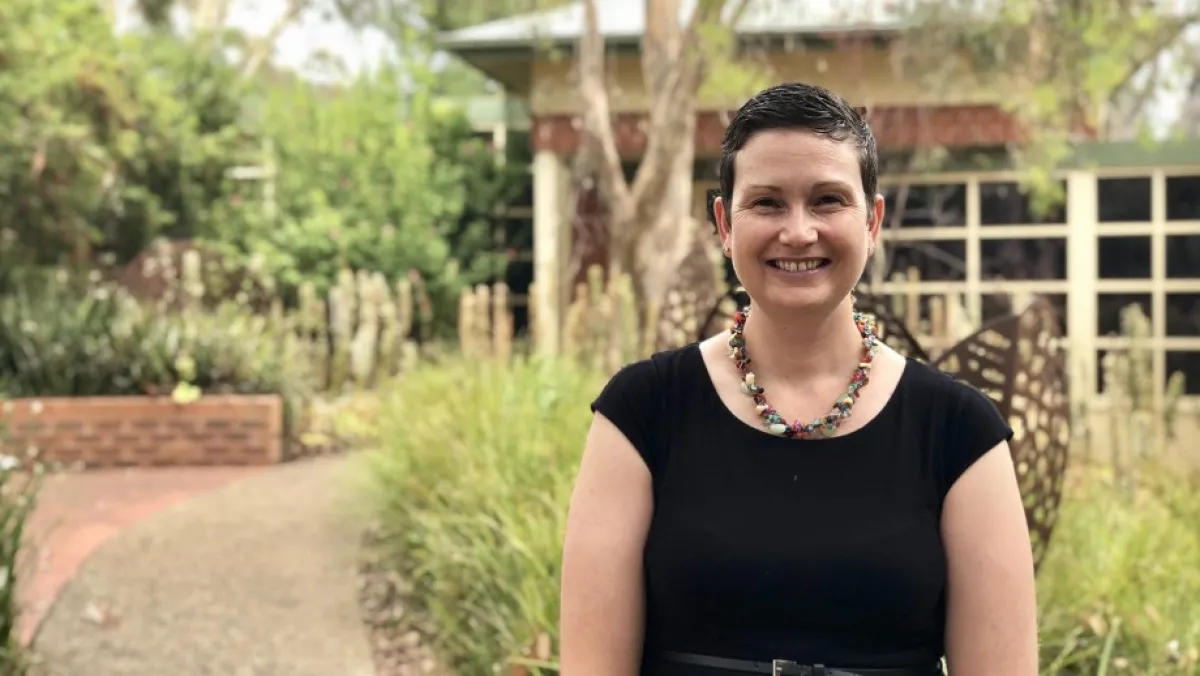 A smiling woman with short hair, wearing a black top and a colorful necklace, stands in front of a lush garden path.