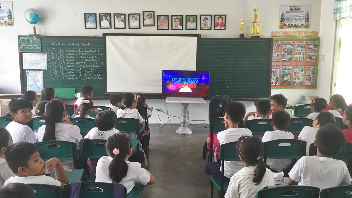 A classroom of students in uniforms paying attention to a video presentation on a television screen at the front of the room.