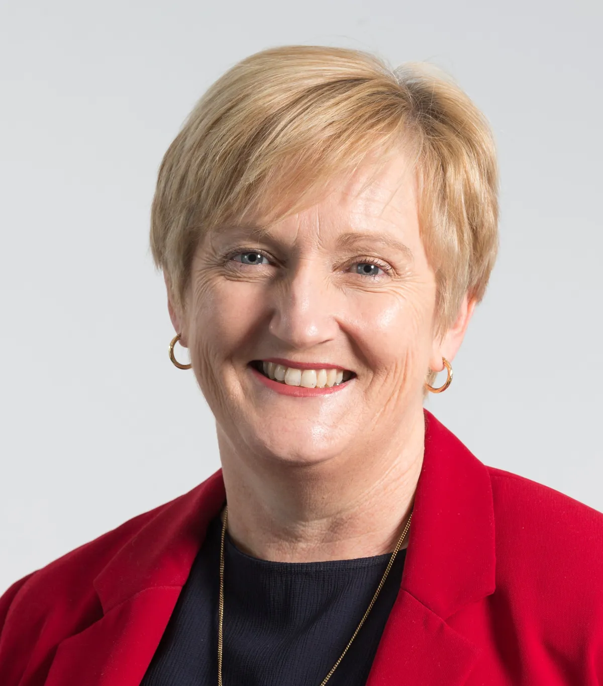 A smiling woman with short blonde hair, wearing a red blazer and gold hoop earrings.