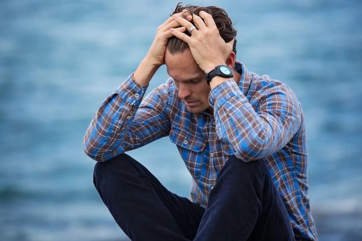 A man in a plaid shirt sits by a body of water, holding his head in distress.