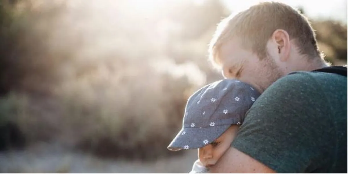 A man tenderly holds a sleeping child against his shoulder in a sunlit outdoor setting.