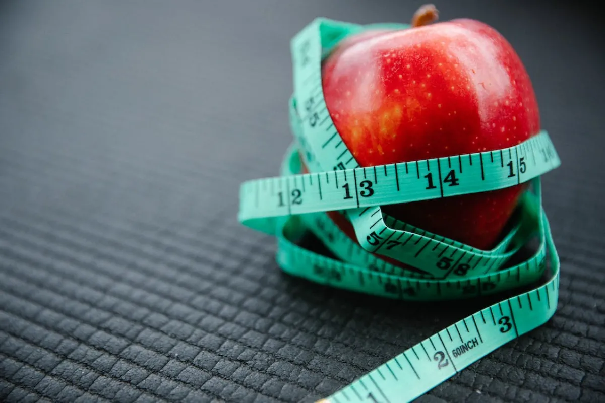 A red apple tightly wrapped with a green measuring tape on a textured dark background.
