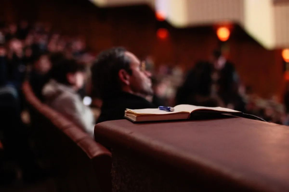 Audience members sitting in a dimly lit theater with a focus on an open notebook resting on an armrest.
