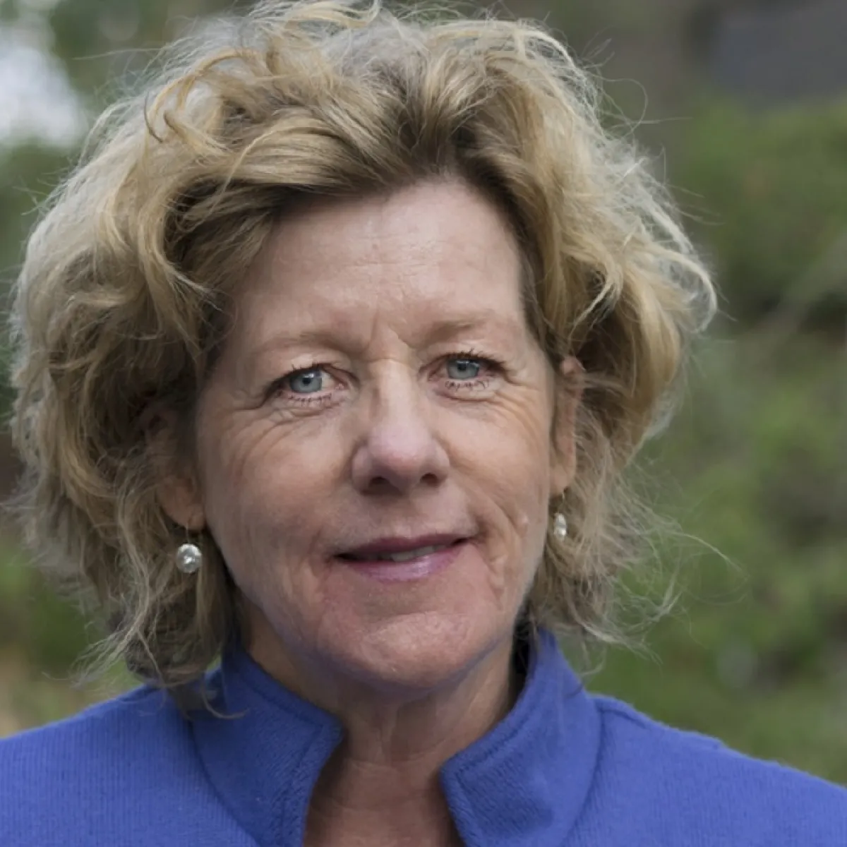 Portrait of a middle-aged woman with tousled blonde hair, wearing a blue turtleneck and small earrings, smiling at the camera.