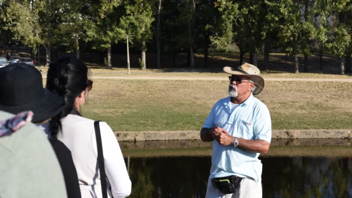 A man in a cowboy hat explains something to a group by the water's edge in a park.