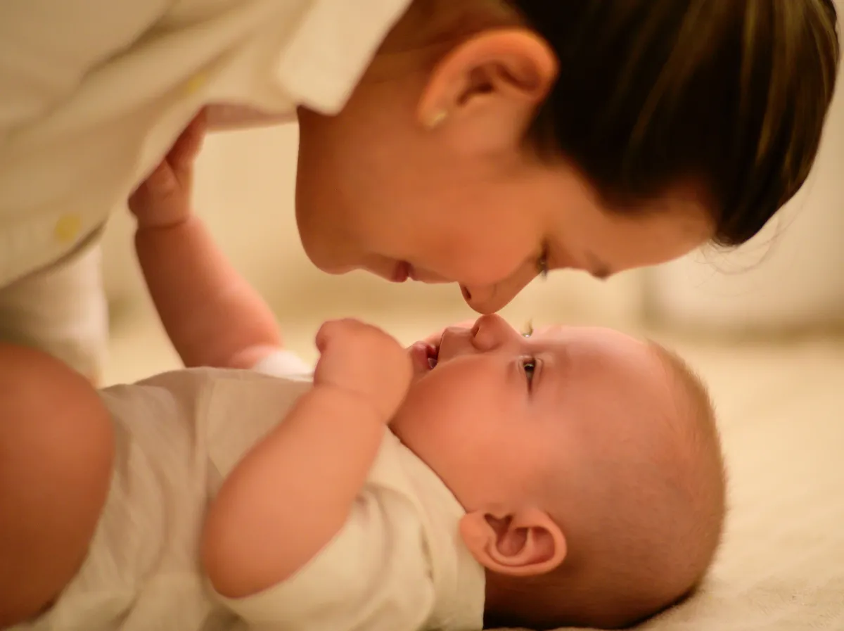 A woman gently touching noses with a smiling baby lying on a soft surface.