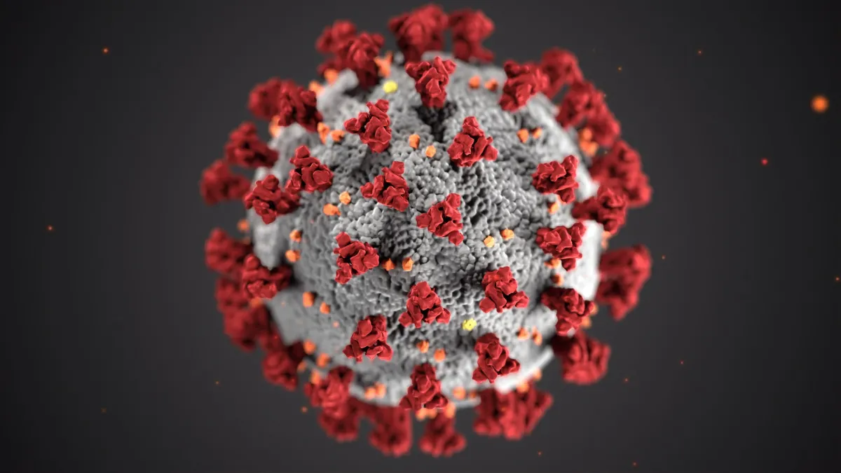 3D illustration of a coronavirus particle with prominent red spike proteins on a grey surface against a dark background.