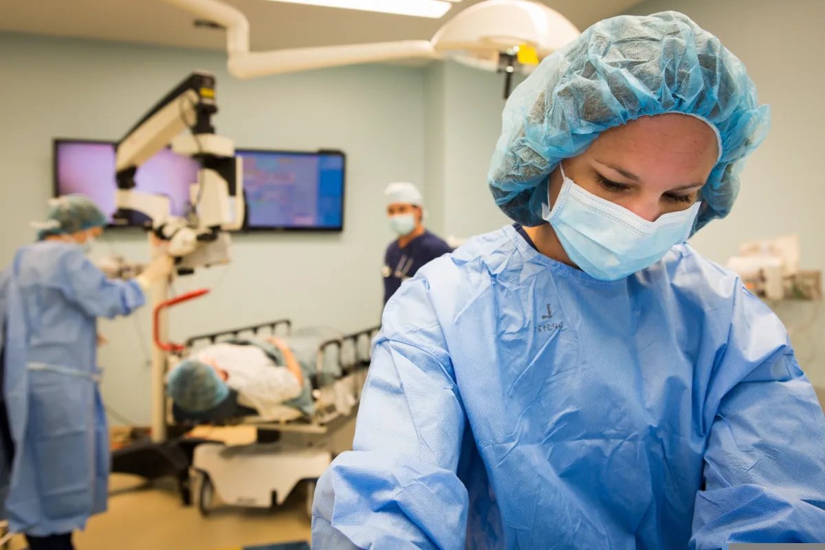 A female surgeon in blue scrubs focuses on her work in a hospital operating room while other medical staff assist in the background.