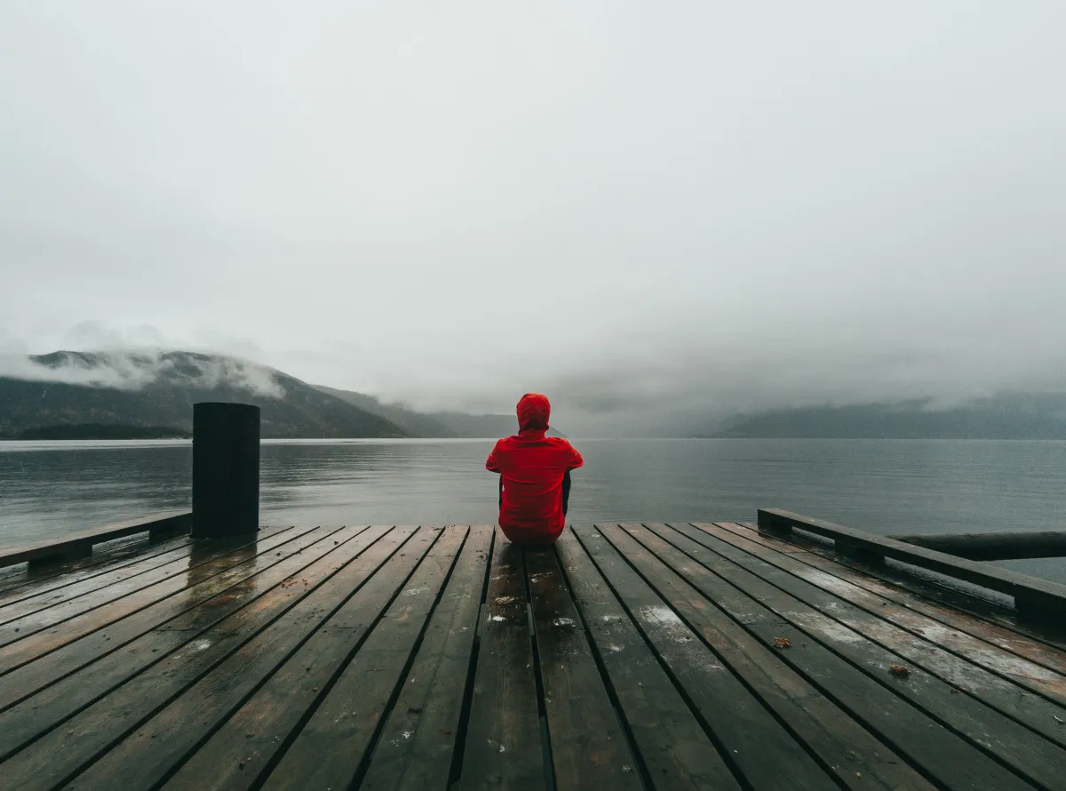 A person in a red hoodie sits at the end of a wooden dock, looking out at a foggy mountain lake.