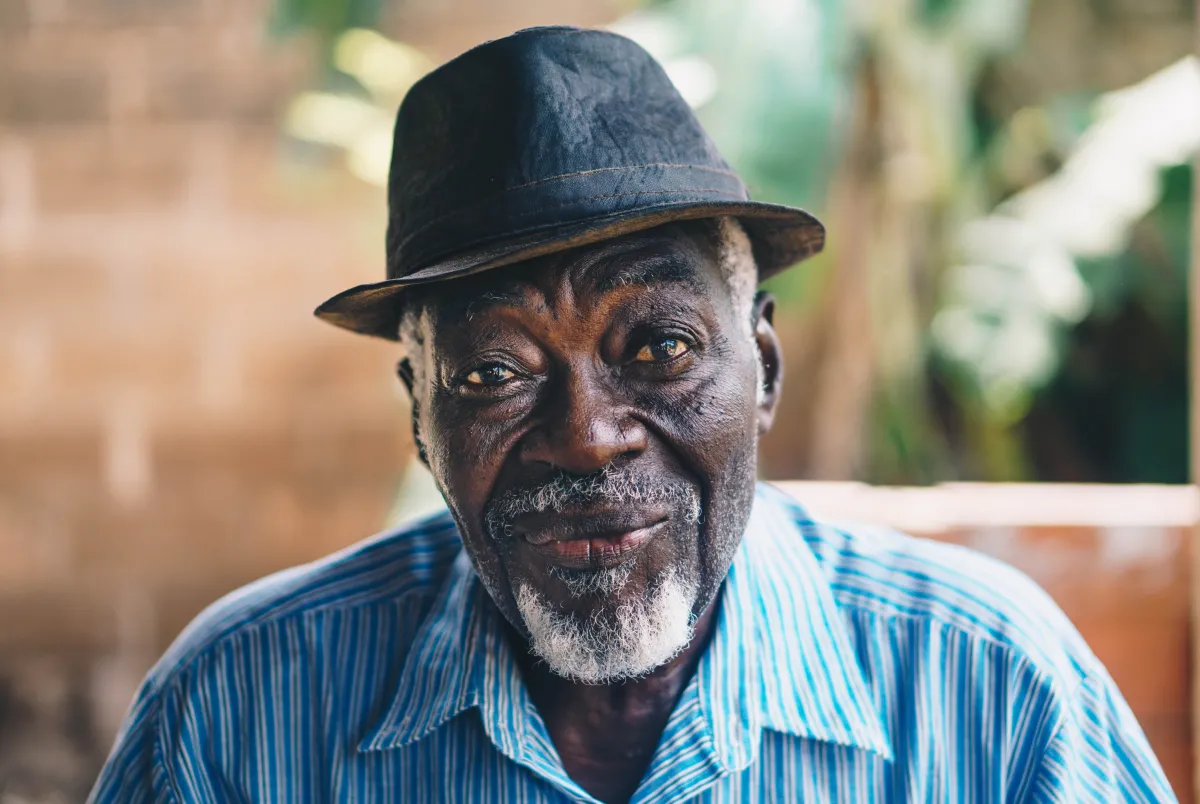 An elderly man with a warm smile wearing a black hat and a blue striped shirt.