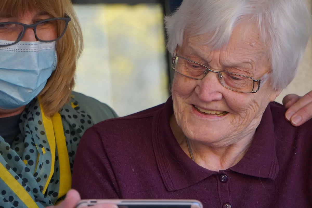 An elderly woman with glasses smiles joyfully while looking at a smartphone, accompanied by another woman wearing a face mask.