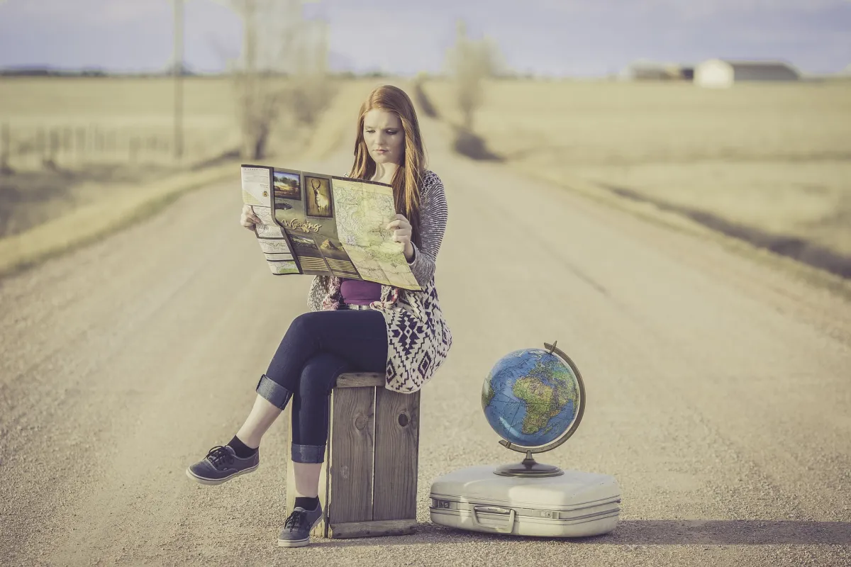 A young woman sits on a suitcase on a rural road, studying a map, with a small globe next to her on another suitcase.