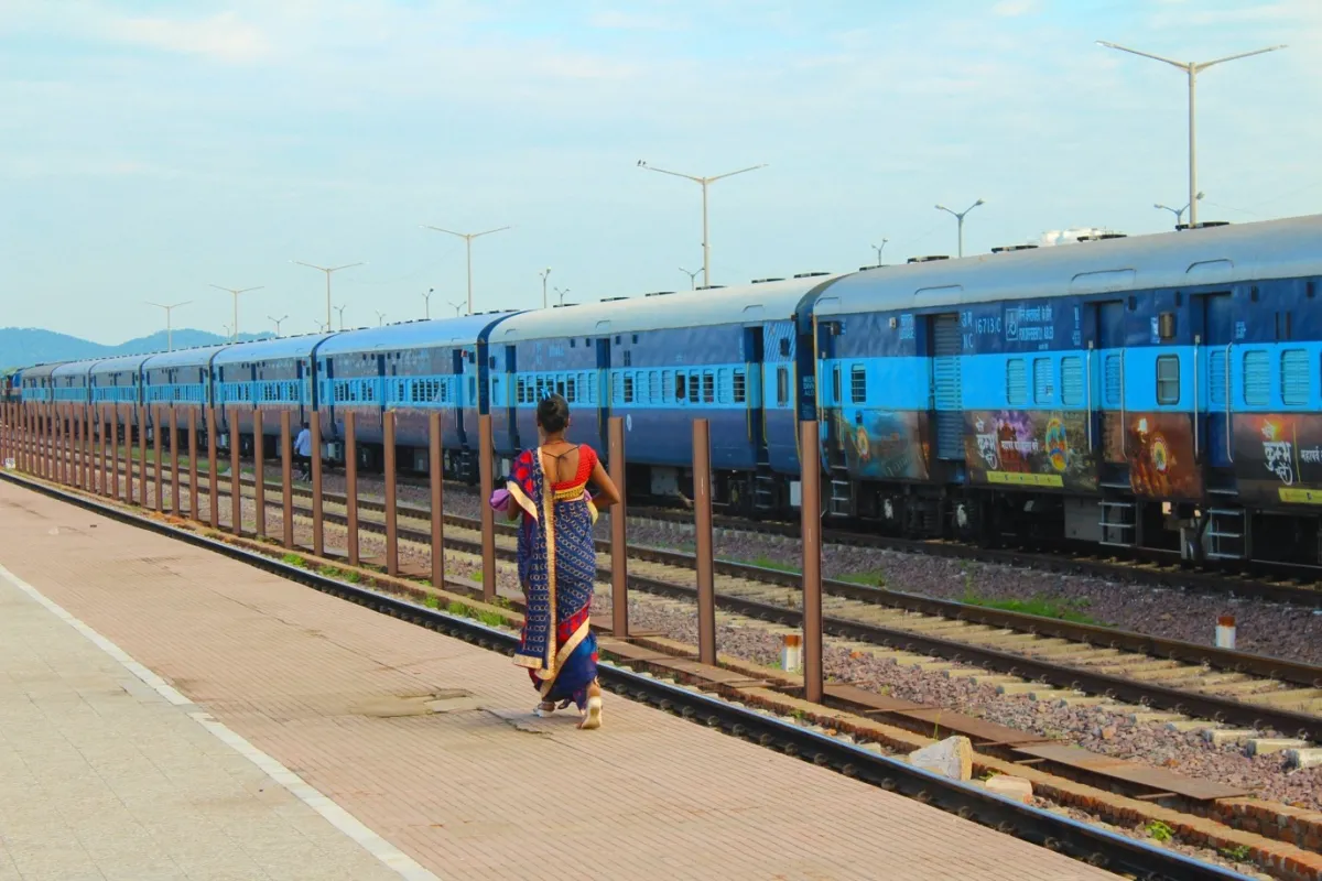 A woman in a sari walks along a platform beside a long, blue train stationed under a clear sky.