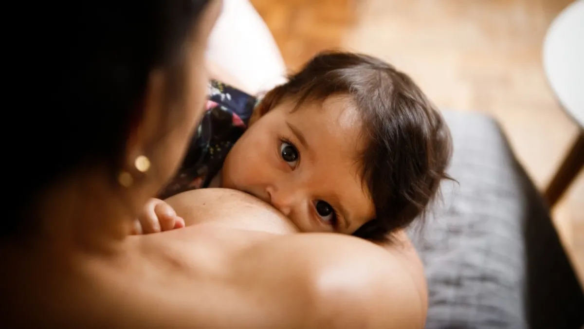 A baby gazes at the camera while breastfeeding from their mother.
